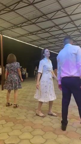 A group of people are gathered under a covered outdoor area with a metal roof supported by beams. The ground is paved with interlocking square tiles in shades of red, beige, and gray. In the foreground, a man in a white dress shirt and black pants stands with his back to the camera. To his left, a woman in a white dress with a floral pattern walks away from him. In the background, to the right, a backdrop decorated with blue and white balloons, streamers, and a "1" signifies a birthday celebration. Several people are standing and interacting around this backdrop, including a person in a bright pink dress with a bow in their hair. The lighting appears to be artificial, with overhead lights and a bright vertical strip light visible. There are also a few chairs and tables set up in the background.