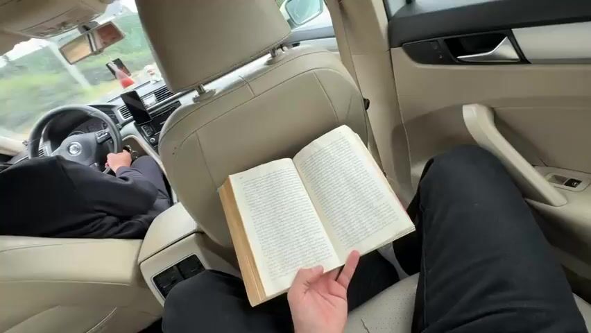 The video captures the interior of a car driving through Tsitsamuri, Georgia, from the perspective of a rear-seat passenger. An individual is seated in the back, holding an open book with visible text on its pages, apparently reading. In the front, another person is driving the car, mostly visible from behind, gripping the steering wheel of a Volkswagen vehicle.

The car's interior features light beige seats, a headrest, and a dashboard. The center console includes an infotainment screen displaying "11:41 PM," indicating the time of day. A smartphone is mounted on the dashboard, and a small, light-colored decorative figurine sits near the windshield. The driver, reflected briefly in the rearview mirror, appears to be wearing glasses.

Outside the car window, a road with a barrier and a green landscape of trees and bushes can be seen. A small structure is visible in the distance, suggesting a highway or main road environment. The overall scene depicts a nighttime journey, with a passenger engaged in reading.