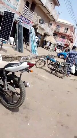The video shows a street scene in a neighborhood in what appears to be a city in Africa. The camera pans across the street, showing buildings, motorcycles, cars, and people walking. In the first part of the video, the camera focuses on two parked motorcycles, one mostly white and the other a faded blue. There are businesses nearby, including one called "GLOBAL SPRING." The camera continues panning to the right, showing people walking on the sidewalks and street. There are parked cars and other motorcycles in the background. Two women wearing traditional Islamic clothing cross the street. A man stands near a building, looking at his phone. The overall scene is a typical street view in an urban African neighborhood.