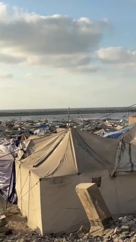 The video shows an aerial view of a densely packed tent city, possibly a refugee camp, stretching towards the sea. The tents are made of various materials, including canvas and tarpaulin, in shades of white, beige, and blue, some appearing worn and damaged. The ground is sandy and strewn with debris, including rocks and pieces of concrete. In the foreground, a larger tent with a peaked roof is prominent, with blue tarpaulin draped over a nearby structure. In the distance, the sea is visible, with a breakwater or pier structure extending into it, and beyond that, a faint line of buildings. The sky is partly cloudy with a light blue hue, suggesting it is daytime. A few people can be seen in the distance, some walking and others tending to the tents, but they are too far away to discern details. The overall impression is one of a makeshift settlement in a coastal area.