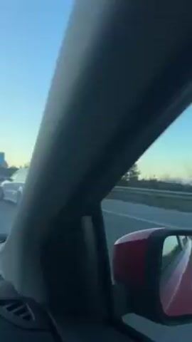 The video, shot from inside a moving vehicle, showcases a white car with extensive aftermarket modifications, including spoilers, body kits, and fender flares. The modified car drives down a highway alongside the camera car. The surroundings include clear skies, distant hills, roadside trees, and a billboard featuring an ice cream advertisement.