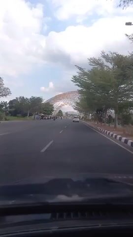 A first-person perspective video shows a road with vehicles and trees. In the distance, a large, arched structure with a geodesic dome-like appearance is visible. The sky is cloudy. The perspective suggests the video is being recorded from inside a vehicle.