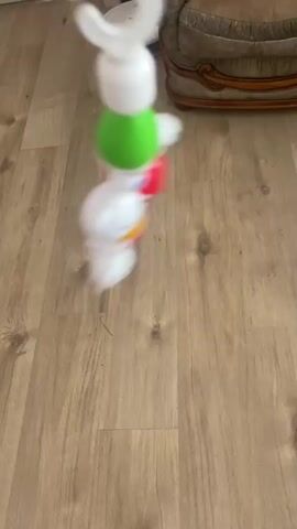 The video begins with a close-up of a toy mobile hanging from a hook. The mobile consists of several brightly colored plastic pieces, including green and white beads, and white figures with red and yellow accents that appear to be characters. The mobile is in motion, causing some parts to be blurred. In the background, a portion of a wooden floor is visible. Further in the background, a robotic vacuum cleaner can be seen on the floor, and the arm of a chair with a patterned fabric is partially visible. The lighting suggests an indoor environment. The video is very short, lasting only a couple of seconds.