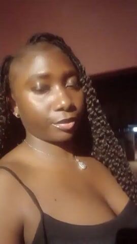 A woman is standing outdoors at night, with a red carpet visible in the background. She is facing the camera and speaking. She is wearing a black top with spaghetti straps and a silver necklace with a pendant. Her hair is styled in long, dark braids. Her complexion is dark and glossy. The lighting is poor, and the background is blurry.