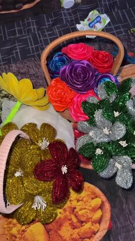 The video shows a still life arrangement of craft items on a patterned surface. In the foreground, there are several artificial flowers made of glittery material in gold, red, and dark pink. A pink woven headband is visible in front of these flowers. Behind these, to the left, is a large yellow artificial flower. To the right, a woven basket is filled with colorful satin rosettes in shades of pink, red, purple, and blue. Adjacent to this basket is a bouquet of artificial flowers in silver and green glitter. In the background, partially visible, are what appear to be food items or decorative objects. There is also a small white container and a wrapper with writing on it. The overall scene is static, with no movement or people visible.