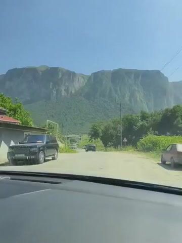 This video captures a point-of-view shot from inside a vehicle, looking forward through the windshield. The vehicle is moving along a paved road that winds through a scenic, mountainous landscape. To the left of the road, there's a low stone wall topped with a wooden fence and some vegetation. A black SUV is parked on the left side of the road. Further down the road, two other vehicles are visible, also moving forward. On the right side of the road, there are trees, a traffic sign, and a utility pole with overhead wires. Towering in the background are imposing, green-covered mountains with steep, rocky cliffs. The sky is clear and blue, indicating a sunny day. The overall impression is a peaceful drive through a natural, rural setting.