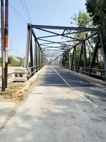 The video shows a first-person perspective of someone driving or walking across a metal truss bridge. The bridge has a paved road with a white line down the center. On either side of the road are metal railings. The bridge is surrounded by trees and vegetation, and the sky is visible above. The bridge appears to be in a rural or semi-rural setting. The camera is moving forward, suggesting progress across the bridge. The lighting indicates it is daytime.