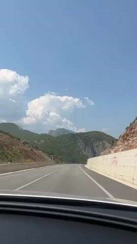 The video shows a first-person view from a car driving on a paved road. The road is surrounded by a hilly, mountainous landscape with green vegetation. The sky is blue with some white clouds. There are other cars on the road. The road has white lane markings and guardrails on the sides. One side of the road has a concrete wall.