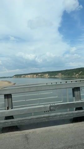 The video shows a view from a bridge over a wide river. The riverbanks are lined with sandy cliffs and green trees. The sky is blue with scattered white clouds. The bridge has a metal railing in the foreground. The video is shot from a moving vehicle, as indicated by the slight movement of the camera.