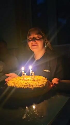 A young woman with blonde hair is in the foreground, smiling at the camera, holding a round cake with two lit candles shaped like the number 23. She wears a black t-shirt with an Adidas logo. The cake is covered in yellow crumbs. The background is dark, suggesting an indoor setting at night, with a hint of light from a window or doorway in the upper right corner. The woman makes a blowing motion with her lips and touches her hair in the latter part of the video.