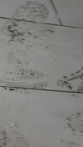 The video shows a tiled floor with numerous dirty footprints. The floor appears to be in a commercial setting, possibly a store, as shelves stocked with products are visible in the background. The footprints are smudged and indistinct, indicating recent activity and possibly a wet or dirty substance on the floor. The lighting suggests an indoor environment. There are no people clearly visible in the frame, but the presence of the footprints implies people have walked through the area. The overall impression is of a floor that has been heavily trafficked and is in need of cleaning.