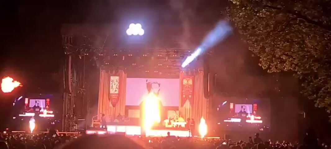 A concert is taking place at night. The stage is lit with bright lights, including spotlights and large screens displaying visuals. Pyrotechnics are set off, creating large bursts of fire on either side of the stage. A crowd of people is visible in the foreground, facing the stage. The overall atmosphere appears to be energetic and festive.