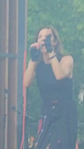 A medium shot video shows a woman with long dark hair wearing a black sleeveless shirt and a black skirt with a wrap in the front. She is on a stage with a wooden backdrop and has on black fingerless gloves. She is holding a microphone with a red cord and appears to be singing. There are microphone stands in the foreground. Smoke or fog is present around the stage.