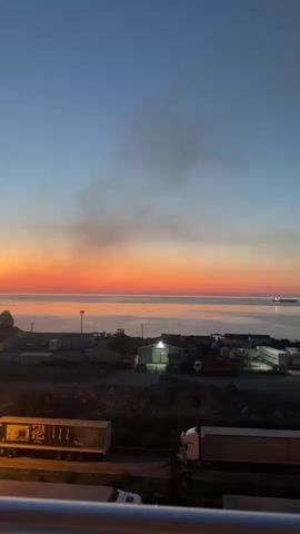 The video shows a view of a body of water at sunset. The sky is a gradient of deep blue at the top, transitioning to orange and red near the horizon. Wisps of smoke are visible in the sky above the water. In the foreground, there is a semi-industrial area with buildings, lights, and several large trucks parked or driving on a road. The water is calm, reflecting the colors of the sunset. A ship can be seen in the distance on the water. The scene appears to be a port or industrial area by the sea at dusk.