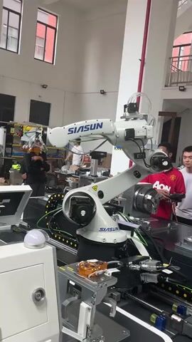 A white industrial robot arm with "SIASUN" branding is actively moving in a well-lit indoor manufacturing or research facility. The robot is positioned over a conveyor belt system and appears to be interacting with an object on a workstation. Several people, including a woman in black to the left and two young men in a red shirt and a grey shirt to the right, are observing the robot's operation. The environment features industrial equipment, shelving units with various items, and exposed ceiling elements like pipes and lights. The overall impression is a demonstration or testing of the robotic system.