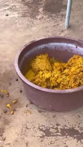 The video shows the process of making palm oil. There's a basket filled with red palm fruits, a basin with mashed yellow palm fruit pulp, and a large metal pot containing a yellow liquid, presumably palm oil. A person is shown using a mortar and pestle to further mash the palm fruit pulp. The ground is earthen and dusty.