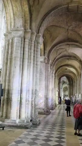 The video pans along a long, arched hallway within a building that appears to be a church or cathedral. The hallway has a checkered floor with large stone columns lining one side. The columns are decorated with carvings and are topped with arches that form the ceiling. There are stained-glass windows visible on the right side of the hallway, casting colorful light onto the walls. Along the right wall, there are several display stands exhibiting colorful fabric art or quilts. Two people are visible in the hallway. One person, wearing a red jacket and a gray skirt, is walking away from the camera down the hallway. Another person, wearing a dark coat and holding a bag, is also walking away from the camera further down the hallway. The overall impression is one of a historic and artistic interior space.