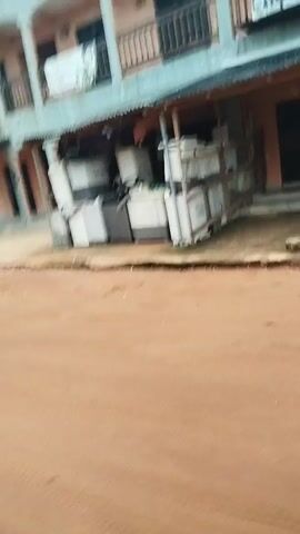 The media file shows a street scene in Isiehor, Nigeria. The foreground is dominated by a reddish-brown dirt road. In the background, a light blue, multi-story building stands with several windows and balconies. Some of the windows have black bars or shutters. There are awnings extending from the building.

To the right of the building, an open-air area displays several large white appliances, possibly washing machines or refrigerators, stacked and arranged. A metal structure, perhaps a stall or shelving unit, is also visible in this area, holding more items. In front of the building, a metal display stand or easel is positioned.

A broken concrete or paved area is visible between the dirt road and the building. There are patches of green grass and some debris scattered on the ground. The sky appears overcast, suggesting a cloudy day.

The image captures a commercial area, likely a marketplace or a shopfront where appliances are sold or displayed. There are no people clearly visible in the media file. The overall impression is one of an active, though perhaps somewhat cluttered, commercial street in a Nigerian town.

There is some text visible on the building, but it is largely illegible due to the image quality and distance. On one of the upper balconies, there appears to be laundry hanging.