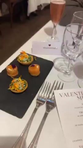 The video shows a table set for a Valentine’s Day dinner, with various glasses, cutlery, and a menu. There are some appetizers on a slate serving board. A name card reads "Mavrommatis." A man with dark hair and a beard is seated at the table, wearing a brown turtleneck sweater. He looks directly at the camera. The setting appears to be a restaurant with other people in the background. The ceiling has exposed wooden beams.