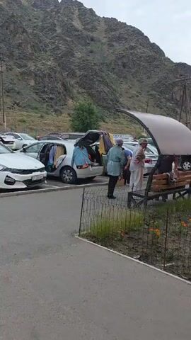 The video shows a parking lot with mountains in the background. Several cars are parked, one of which has its trunk open and clothes spilling out. There are also benches and a covered seating area. A few people are visible, including two in medical scrubs standing near the car with the open trunk, and a family eating at a table under the covered seating area. A young boy walks past the seating area. The scene appears to be in a somewhat rural or undeveloped area, possibly a tourist spot given the seating and the presence of a cafe or restaurant.