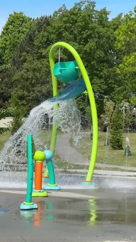 The video shows a water park feature in operation on a sunny day. The main structure consists of a curved lime green pipe supporting a green bucket which tips over when filled with water, splashing onto a blue slide below. The water then runs down and splashes to the ground. Nearby are several shorter, colorful water features, some spraying water upwards. The ground is wet from the water. The park is surrounded by green trees and grass. A paved walkway is visible in the background, and a car drives by.