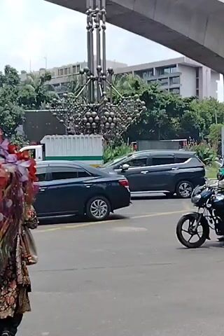The video shows a busy street scene with traffic and pedestrians. In the foreground, a motorcycle with two riders, both wearing helmets, is moving. Behind them, a green auto-rickshaw is also visible. Further back, a white van and a dark-colored car are in traffic. A large metallic sculpture, possibly abstract art, is prominent in the center of the frame, beneath a concrete overpass. The background features buildings and lush green trees. A pedestrian, an elderly man with a white beard and a cap, walks past in the lower left corner. The scene appears to be a daytime urban environment.