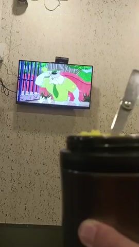 A person's hand is holding a dark brown thermos with a silver metal spoon inside, filled with yellow substance. In the background, a television screen displays a cartoon character in a green shirt and tie. The wall behind the television has a textured, light-colored pattern. A laptop is visible in the lower foreground.