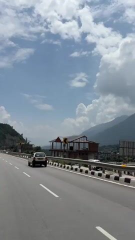 A white car drives on a highway with a mountainous landscape in the background. A building with a modern design and glass windows is situated on the side of the highway. The sky is blue with scattered white clouds. The road has white lines indicating lanes and a guardrail along the side. The overall scene depicts a scenic drive through a hilly region on a cloudy day.