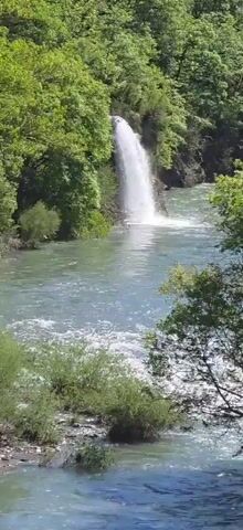 The video shows a river flowing towards the camera, with a waterfall visible in the distance. The river is bordered by green trees and vegetation. The water is a light blue color, and it appears to be moving rapidly. The waterfall is relatively small, and it is surrounded by trees.