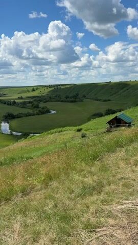 The image depicts a wide, panoramic view of a verdant, rolling landscape under a bright, partly cloudy sky. The foreground is dominated by a grassy hillside, with dry, straw-colored grass interspersed with green blades. A wooden structure with a green roof is nestled on the slope to the right of the frame, overlooking the expansive valley.

In the mid-ground, a sinuous river meanders through a lush green valley. The river's surface reflects the sky and surrounding foliage, creating streaks of light and shadow. Patches of trees and bushes dot the valley floor, adding texture and depth to the scene. The landscape extends into the distance, with rolling hills that fade into a hazy horizon. The overall impression is one of natural beauty and tranquility.

The time of day appears to be daytime, likely late morning or early afternoon, given the bright sunlight casting shadows. The weather seems pleasant, with a mix of sunshine and cumulus clouds. The location is identified as Kamaevo, Russia, which suggests a rural or natural setting. There is no discernible activity or event taking place; the image captures a static, serene landscape. No text is visible in the media file.
