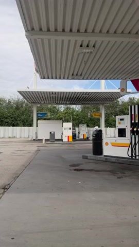 A stationary, eye-level, medium shot captures a deserted gas station under a bright, overcast sky. The foreground is dominated by a concrete forecourt with a few dark patches. A white, corrugated metal roof, supported by thick white pillars, covers the scene. Under the roof, on the right, a Shell V-Power branded gas pump stands, equipped with four nozzles. To the left of the pump, a white car is partially visible. Further left, another gas pump is visible, also branded with Shell. In the background, lush green trees form a dense line. The overall atmosphere is quiet and empty.