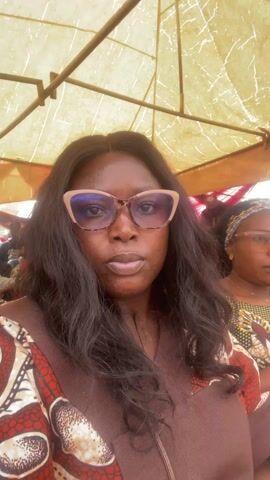 A woman with dark skin, glasses, and long dark hair is recording a selfie video. She is wearing a brown dress with an African print pattern. The background suggests she is under a tent or canopy, with other people seated around her. A woman in glasses and a patterned top is partially visible behind her. The camera occasionally moves slightly, showing more of the surrounding crowd and the canopy structure overhead.