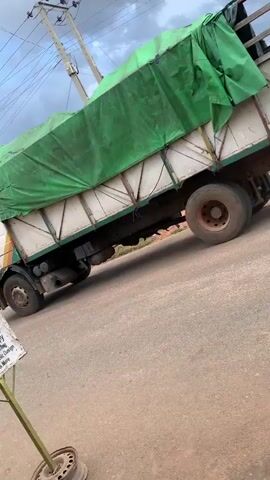The media file captures a scene on a dusty road in Barakin Akawo, Nigeria. A large, white truck with a green tarp covering its cargo drives past. The truck has dual rear wheels and a yellow stripe on its cab. In the foreground, a weathered sign on a metal stand advertises "ELLACOM VENTURES" and lists services like "Computer & Business Centre," "Typing & Photocopy," "General Typing," "Binding," "Lamination," and "Graphic Design." The sign also mentions "Internet Services & More."

The setting appears to be a somewhat rural or semi-urban area, with buildings visible in the background, including one with a green corrugated roof. The road is unpaved and dusty. The sky is partly cloudy with dramatic, dark clouds, suggesting it might be late afternoon or that a storm is approaching. There are power lines overhead.

The activity is simply the truck driving down the road. There are no prominent individuals visible, though a blurred figure might be seen in the distance near some buildings. The overall mood is mundane, depicting everyday life and commerce in the depicted location. The visible text on the sign provides specific details about the services offered by a local business.