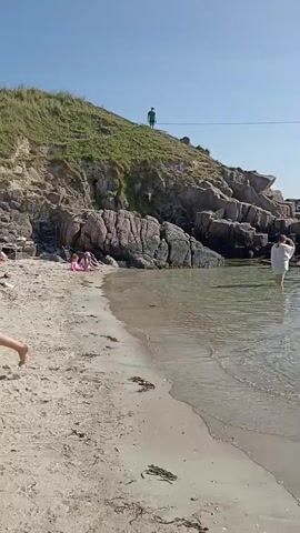 The video shows a scenic view of a beach and a small harbor. In the foreground, there is a sandy beach with some seaweed. The water is clear and shallow near the shore, with gentle waves. In the middle ground, there is a rocky coastline with a few people scattered along it. One person in a white shirt is wading in the water. Further back, a group of people are gathered near a small harbor, where several boats are docked. There are also a few cars parked along the harbor. The background is dominated by a grassy hill with a clear blue sky above. There is a person in a green outfit standing on the grassy hill. The overall impression is a sunny day at a peaceful coastal location.