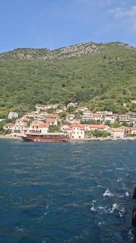 The video shows a body of blue water with waves. In the background, there is a village with white buildings and red roofs nestled at the base of a lush green hill. A large brown boat with multiple decks is docked near the shore. The perspective is from another boat, showing a white and red railing and some people's legs on the right side of the frame. The sky is clear and blue. The camera appears to be moving forward, creating a wake in the water.