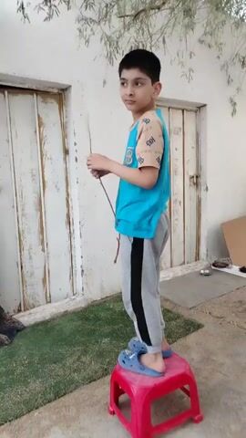 A young boy stands on a red stool, holding a stick in his right hand. He is wearing a blue t-shirt with a tan and brown pattern on the sleeves, gray sweatpants with a black stripe down the side, and blue Crocs. He turns his head to the left and then raises his right arm, reaching towards the branches of a tree. The boy is positioned in front of two white doors. The ground is covered with dirt and some green artificial turf. The environment appears to be outdoors, with a tree overhead and a wall behind the doors.