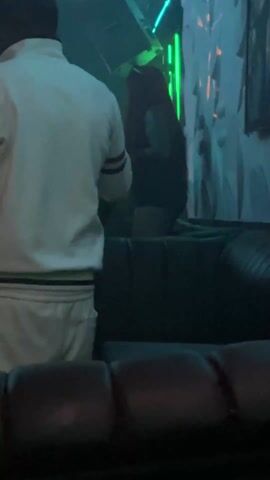 The video shows a dimly lit nightclub scene. A person in a white tracksuit is standing in the foreground, facing away from the camera. To their right, a woman with long, reddish-orange hair and wearing a dark top and shorts is dancing on top of a large, dark leather sofa. The sofa is positioned against a wall decorated with textured patterns. The lighting in the club shifts between shades of blue and green, with some overhead fixtures visible. The audio suggests a lively atmosphere with music playing.