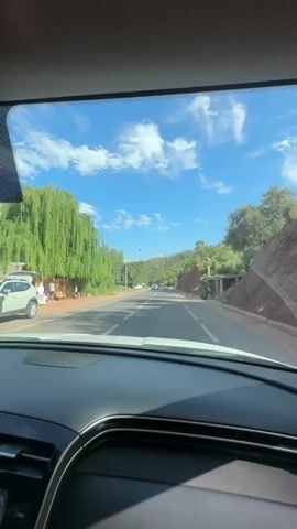 The view is from the driver's seat of a car driving down a two-lane road surrounded by trees and mountains. The sky is blue with white clouds. Other cars are visible on the road ahead. A person is seen walking on the left side of the road. The dashboard of the car is visible. The car is moving forward.