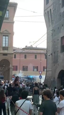 The video depicts a street scene in Bologna, Italy, with a crowd of people gathered. In the background, police vehicles are present, and there is evidence of some form of dispersal or crowd control activity, possibly involving tear gas or smoke, indicated by plumes of white substance. The overall atmosphere appears tense, with a mix of concern and observation from the onlookers.

The subjects include a large group of diverse individuals, some appearing to be protesters or spectators, and several police vehicles. Objects of note include street furniture, buildings with traditional Italian architecture, and what appear to be security barriers or temporary structures. The setting is a narrow urban street lined with historic buildings. One building on the left has arches and a pale facade, while the building on the right is a tall, dark brick structure. In the distance, a pink building with multiple windows is visible.

The activity taking place is a gathering of people in the street, with a visible police presence and signs of an event involving crowd management. The time of day appears to be daytime, with bright, possibly hazy, sunlight suggesting it could be late afternoon or early evening. The weather is unclear, but the presence of smoke suggests potential weather conditions that might exacerbate its effects.

Notable details include a woman standing on a raised surface in the foreground, observing the scene. A traffic sign indicating a right turn is visible near the police vehicles. The emotions conveyed are difficult to ascertain definitively from the still images, but the crowd's density and the police presence suggest a situation that is at least noteworthy and potentially confrontational. There is no visible text that can be read clearly in the provided frames.