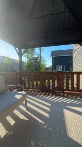 The video shows a sunny backyard patio with a gazebo. A cream-colored outdoor sofa is positioned on the left side of the frame. The sun casts strong diagonal shadows from the gazebo onto the beige outdoor rug covering the patio floor. In the background, a wooden fence encloses the yard, and a dark blue house is visible to the right. Bubbles float in the air near the bottom right of the frame. The camera appears to be at a low angle, possibly from someone sitting or lying on the sofa. The overall atmosphere is peaceful and sunny.