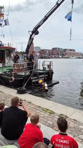 A group of onlookers, mostly men and boys, are seated on a wooden bench by a waterfront. They are watching a dive team operating from a boat. One diver, wearing a full diving suit and helmet, is being lowered into the water by a crane mounted on the boat. Two men in uniform are on the boat, one appears to be operating the crane and the other is talking. A flag is attached to the crane. The boat has the text "Taucher- u. Bergungsgesellschaft Bremen" on its side. The background shows a harbor with buildings and other boats under a cloudy sky.