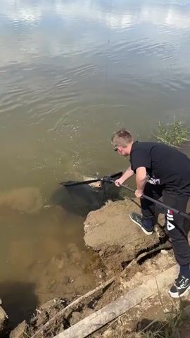 A young boy in sunglasses and a black t-shirt is fishing by a muddy riverbank. He is holding a large fishing net with a long handle, lifting it out of the water. A fish is visible in the net, and the boy appears to be in the process of landing it. The water is murky, and the riverbank is eroded, with some debris visible. The sky is overcast, with reflections on the water.