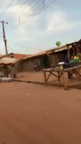 The video shows a street scene in a rural or developing area. The road is unpaved, reddish-brown dirt. A motorcycle drives past a market area with stalls displaying fruits and vegetables. Several individuals are present; some appear to be vendors or shoppers. Buildings in the background are simple structures, some with corrugated metal roofs. Overhead are power lines and communication towers. The sky is overcast, suggesting either early morning or late afternoon.