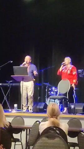 A man and a woman stand on a stage in front of a black curtain. The man is on the left, wearing a blue shirt with intricate embroidery and beige pants. He holds a microphone and stands behind a podium with a laptop on it. The woman is on the right, wearing a red shirt with embroidery and black pants. She holds a microphone to her mouth. Behind them, a drum set is visible, along with other stage equipment. The stage is lit from above, with some lights casting a green glow. In the foreground, the backs of several people are visible, seated at tables, with their heads and shoulders in frame. The overall environment appears to be an indoor performance or presentation space.