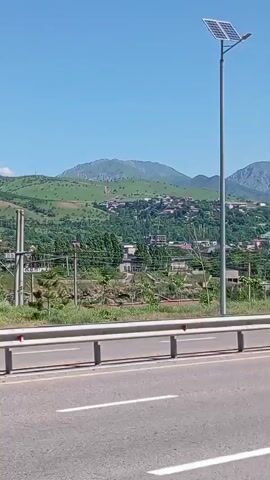 The camera is moving on a road with a gray metal guardrail beside it. Beyond the guardrail, the landscape is lush green with rolling hills and mountains in the background. The sky is clear and blue. Utility poles and lines are visible in the distance.  The houses can be seen scattered in the landscape. An ISUZU truck passes by on the road.