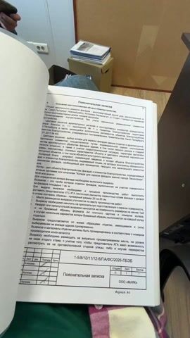 The video shows a person flipping through pages of a large document with various diagrams and text written in Russian. The document appears to be a technical or official report of some kind. The person is wearing a blue and white plaid shirt and has a ring on their finger. In the background, there is a desk, a cardboard box, and another person holding a mobile phone. The setting seems to be an office environment. The actions are limited to turning pages of the document.