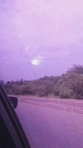 This is a video of a roadside view, likely from inside a vehicle. The scene is dominated by a road on the right side, bordered by dense green foliage and trees. The sky is a hazy purple, suggesting either a sunrise, sunset, or unusual atmospheric conditions like fog or pollution. A geometric, possibly man-made structure, is visible in the distance, reflected in what appears to be a window or glass surface in the foreground. The purple hue extends across the entire frame, creating a surreal and slightly mysterious atmosphere. There are no people clearly visible, but a portion of a dark vehicle's interior is in the lower right corner. The overall impression is one of travel, possibly on a rural road in Dukku, Nigeria, under a peculiar sky. The time of day is ambiguous due to the lighting.