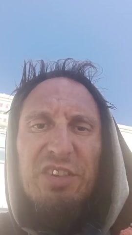 The video shows a close-up of a man's face. He has dark hair, a goatee, and is wearing a hooded sweatshirt. The background is a clear blue sky with the top of a building visible. The man is speaking to the camera.
