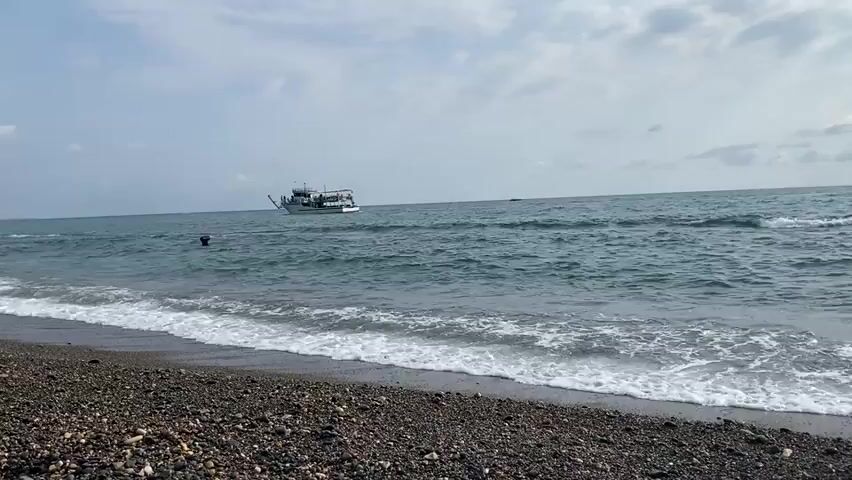 The video captures a scene from a pebble beach looking out towards a moderately wavy ocean. A white boat with a shaded upper deck, carrying several passengers, sails across the water in the distance. The sky is overcast with scattered clouds. In the foreground, waves break on the shore, creating white foam. A dark object, possibly a buoy or debris, is visible in the water closer to the shore.