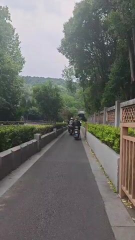 A video shows a person on a scooter driving down a paved path. The path is bordered by low hedges on the left, and a decorative fence and additional hedges on the right. The scooter drives towards a tunnel or overpass visible in the distance, with green hills beyond. The sky is overcast. The video seems to be taken from a moving perspective, likely by another person following behind.